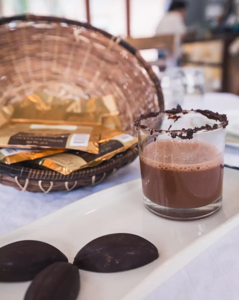 A glass of chocolate drink with a chocolate-coated rim sits on a white tray beside three pieces of dark chocolate; in the background, a basket filled with Bohol gold-wrapped chocolate bars is visible.