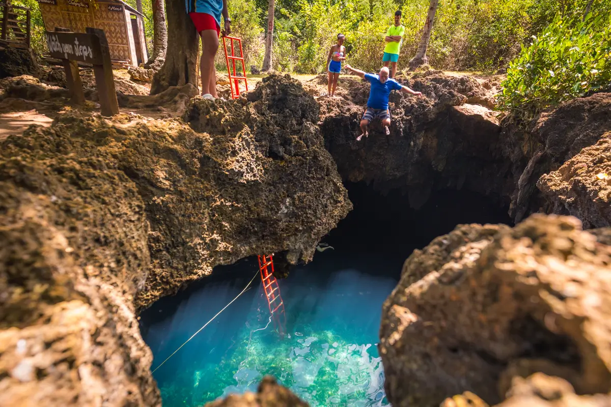 A person jumps from a rocky ledge into a clear blue natural pool below in Bohol, while three others stand nearby. A red ladder descends into the water, surrounded by trees and rugged rocks.