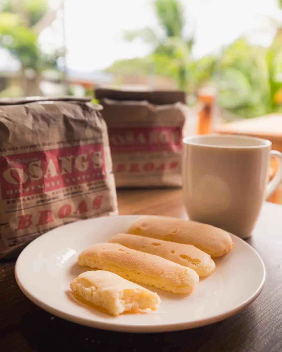 A white plate with four pieces of Bohol broas (ladyfinger biscuits), one partially eaten, sits next to a white mug filled with a hot drink. Bags labeled Osangs Broas are in the background, with greenery outside.