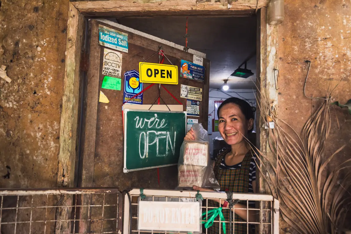 A woman stands behind a counter in a rustic Bohol shop, smiling and holding a plastic bag. Signs on the wall read OPEN and were OPEN, with various stickers and notices visible around the window frame.
