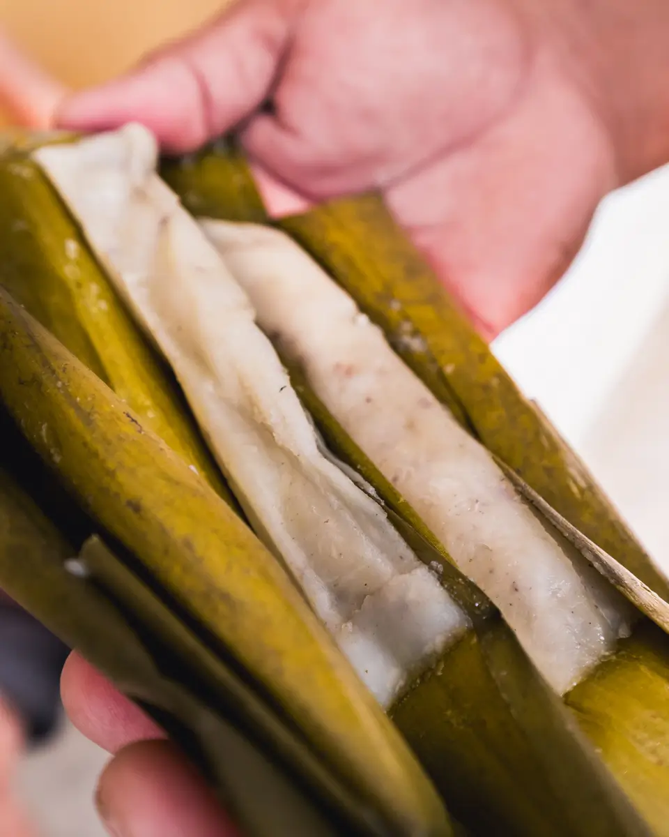 A hand holds an opened banana leaf wrapping, revealing a strip of white, sticky rice cake inside—a popular delicacy from Bohol. The rice cake has a smooth texture and is partially covered by the leaf.
