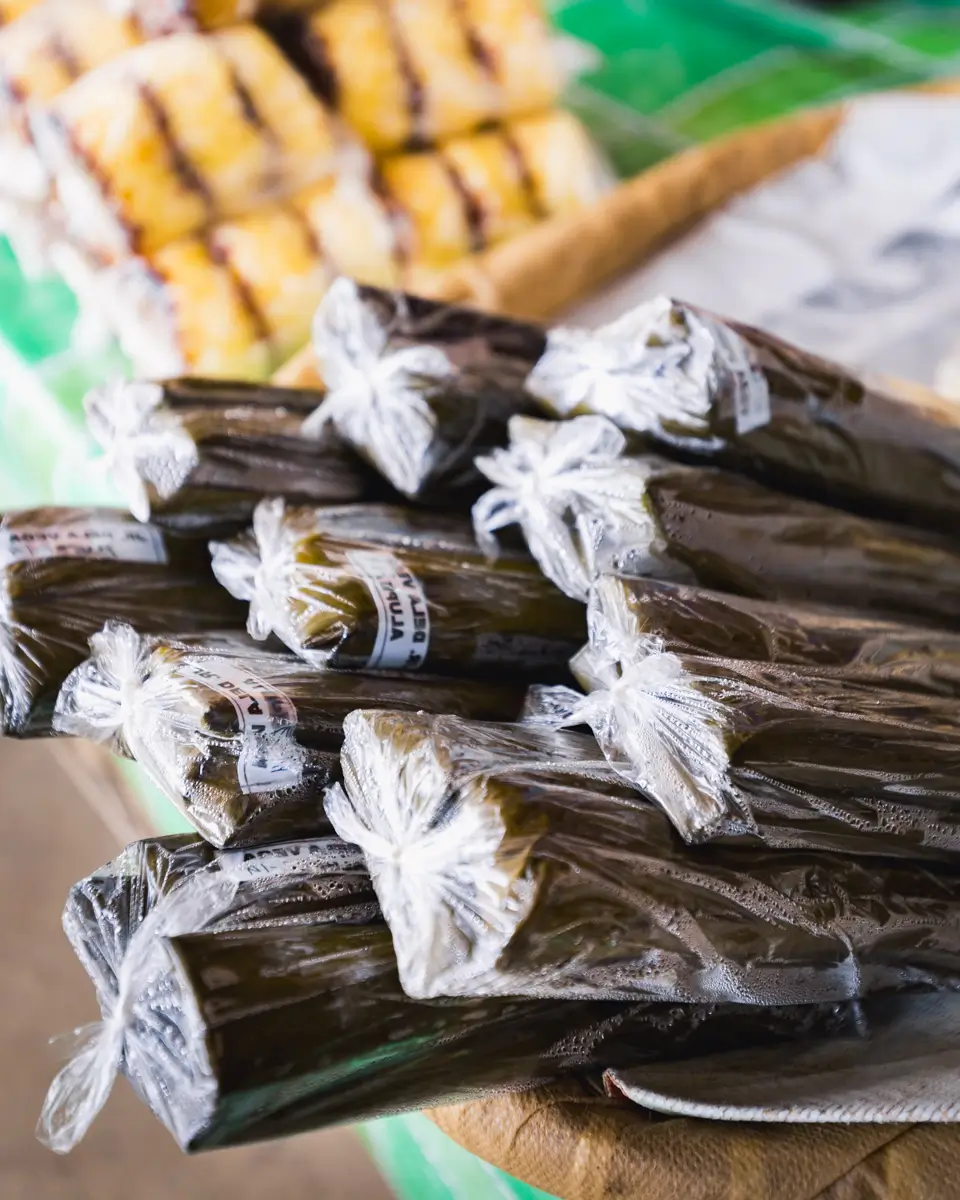 A close-up of several pieces of Bohol delicacies wrapped in banana leaves and tied with plastic, stacked together at a market stall. More wrapped items are visible in the blurred background.