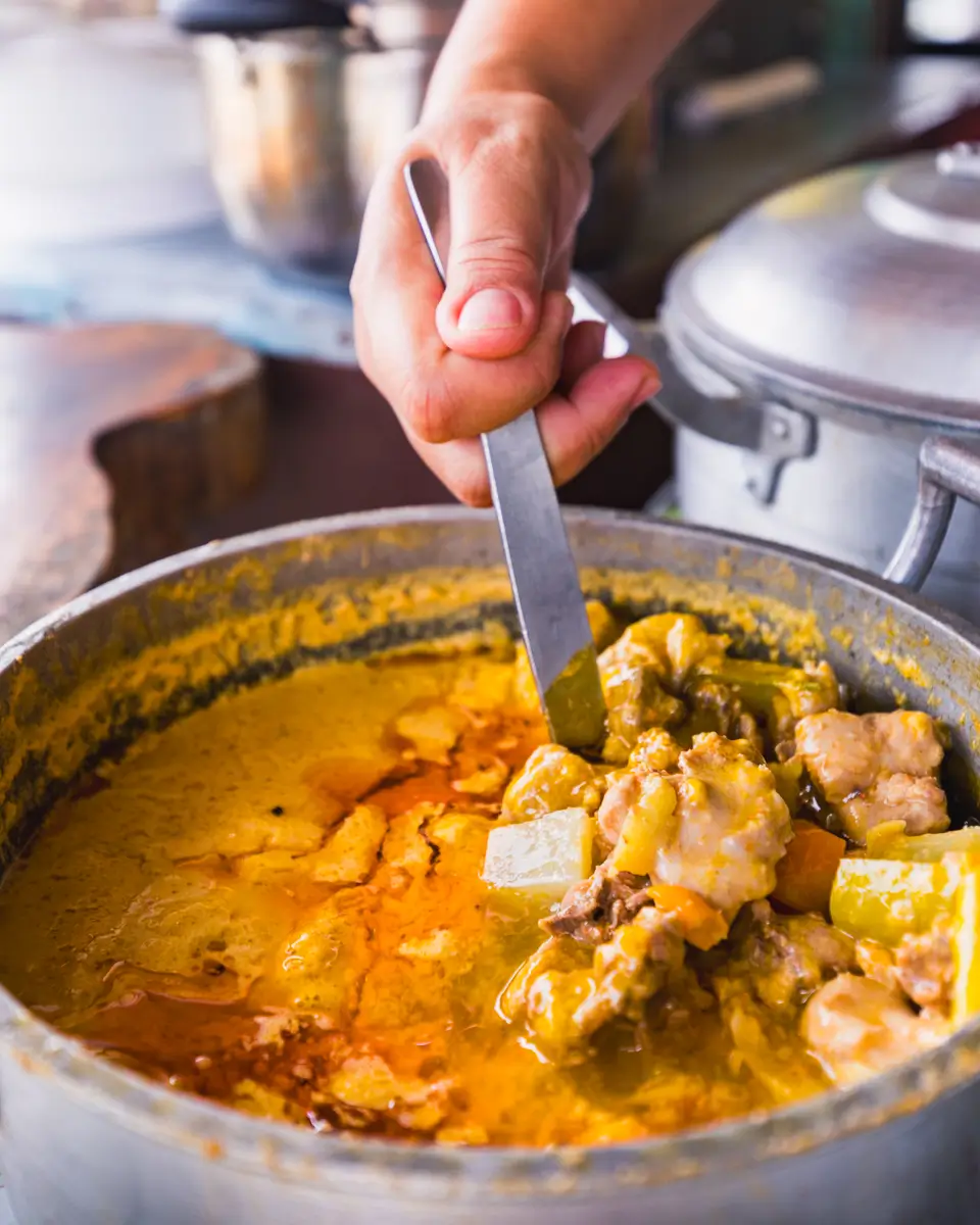 A hand holds a metal ladle, stirring a pot of thick, oily yellow curry with chunks of meat and tofu—a savory dish inspired by Bohol. A pot lid and wooden cutting board are visible in the background.