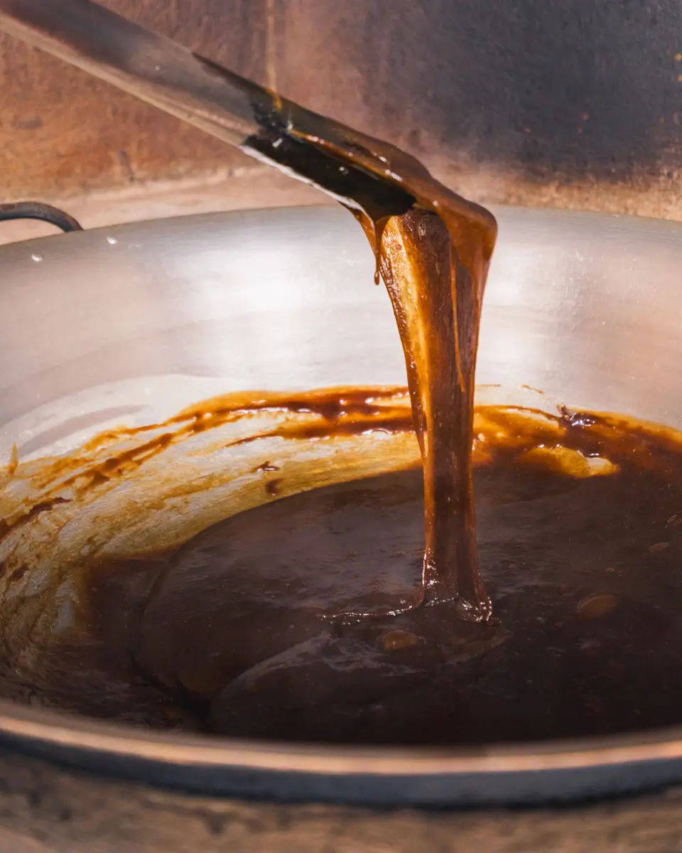 A thick, dark brown syrup, possibly a Bohol delicacy, is being stirred in a large metal pan with a wooden spatula. The mixture appears glossy and viscous, likely being cooked or reduced over heat.