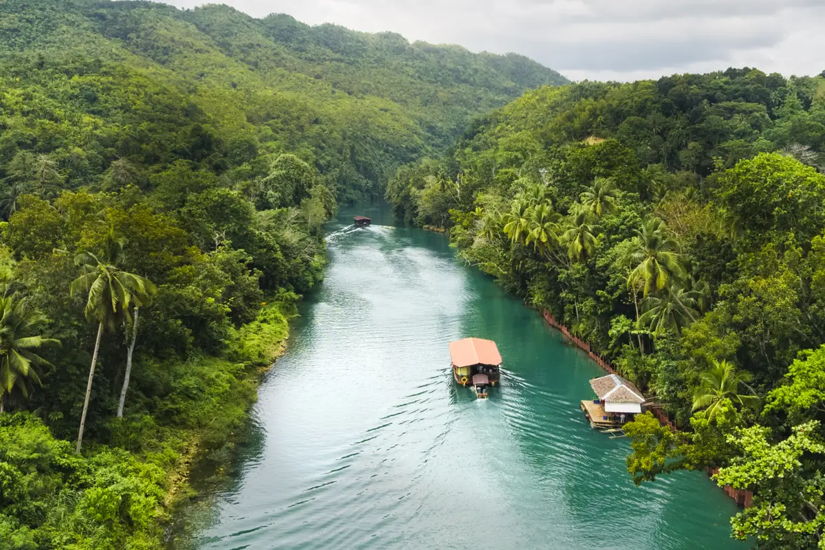 A wide river winds through Bohol’s dense green forest, with a few boats—including a floating house and a larger boat with a roof—cruising along the water under cloudy skies. Hills covered in trees rise in the background.