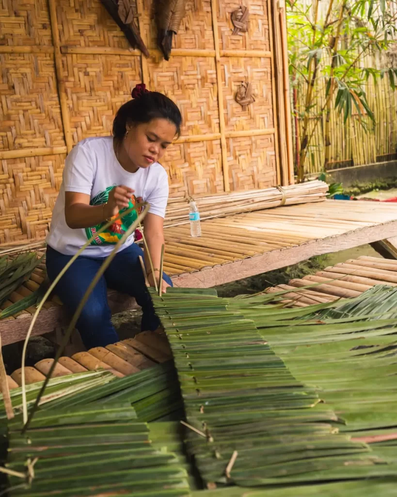 A woman in a white shirt sits on a wooden platform in Bohol, weaving green palm leaves. She is outdoors near a bamboo wall, surrounded by woven bamboo and lush foliage. A plastic water bottle rests beside her.