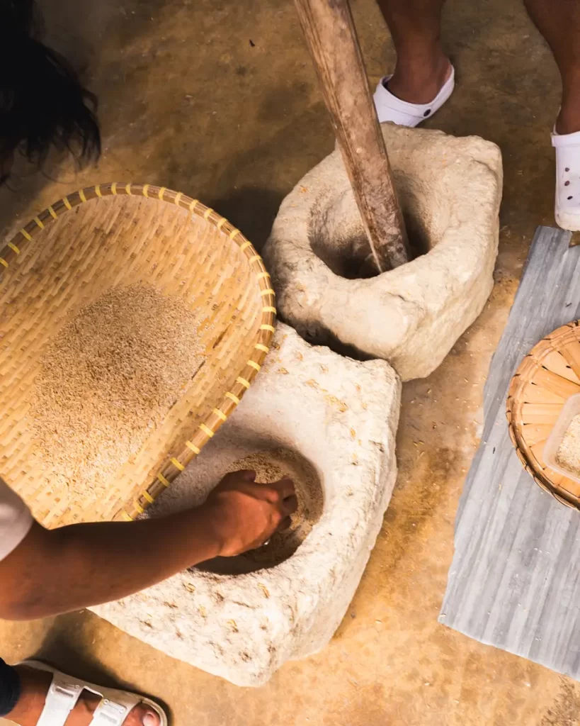 A person in Bohol uses a traditional stone mortar and pestle to grind grains beside a woven basket of processed grains on a brown floor. Only hands, feet, and part of the mortar are visible.
