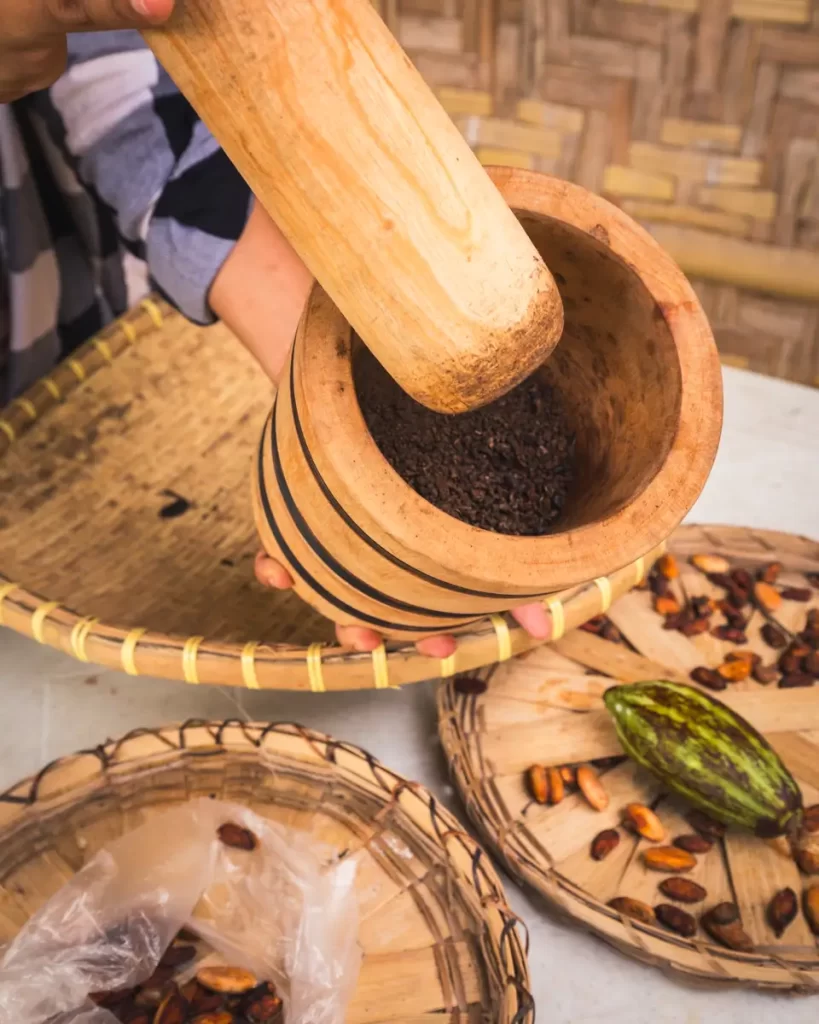 A person uses a wooden pestle to grind cacao beans in a wooden mortar in Bohol. Nearby, cacao beans and a cacao pod rest on woven trays set on a white surface.