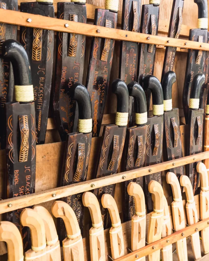 Close-up view of several traditional Bohol wooden machete sheaths with decorative carvings, arranged vertically in rows on a display rack, with wooden-handled machetes below them.