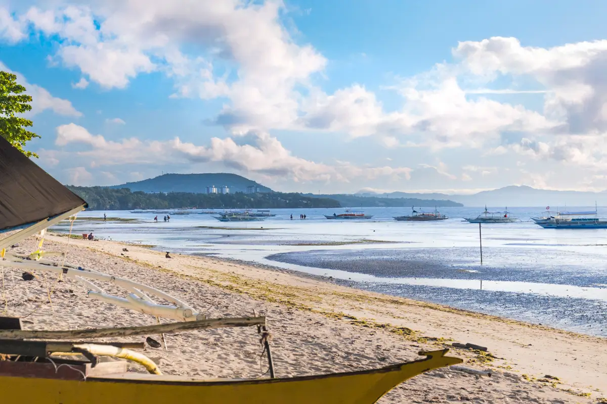 A sandy beach in Bohol with shallow water at low tide, several boats anchored near the shore, distant hills on the horizon, and scattered clouds in a blue sky.