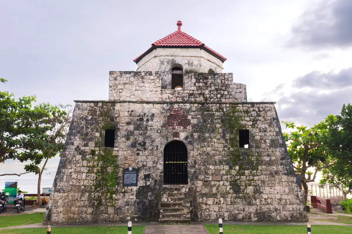 A stone fort in Bohol with a red-roofed watchtower, rectangular base, arched doorways, and small windows stands on grass with trees and the sea in the background. The sky is cloudy.