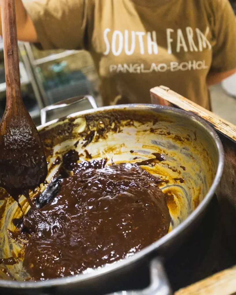 A person stirs a thick, dark mixture in a metal pot with a wooden spoon. Wearing a brown shirt that reads “SOUTH FARM PANGLAO-BOHOL,” they showcase traditional cooking techniques from Bohol.