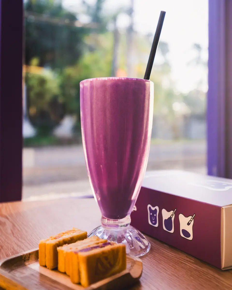 A tall glass of purple smoothie with a black straw sits on a wooden table in Bohol, next to a small wooden tray with sliced cake and a purple box featuring white drink and cat icons.