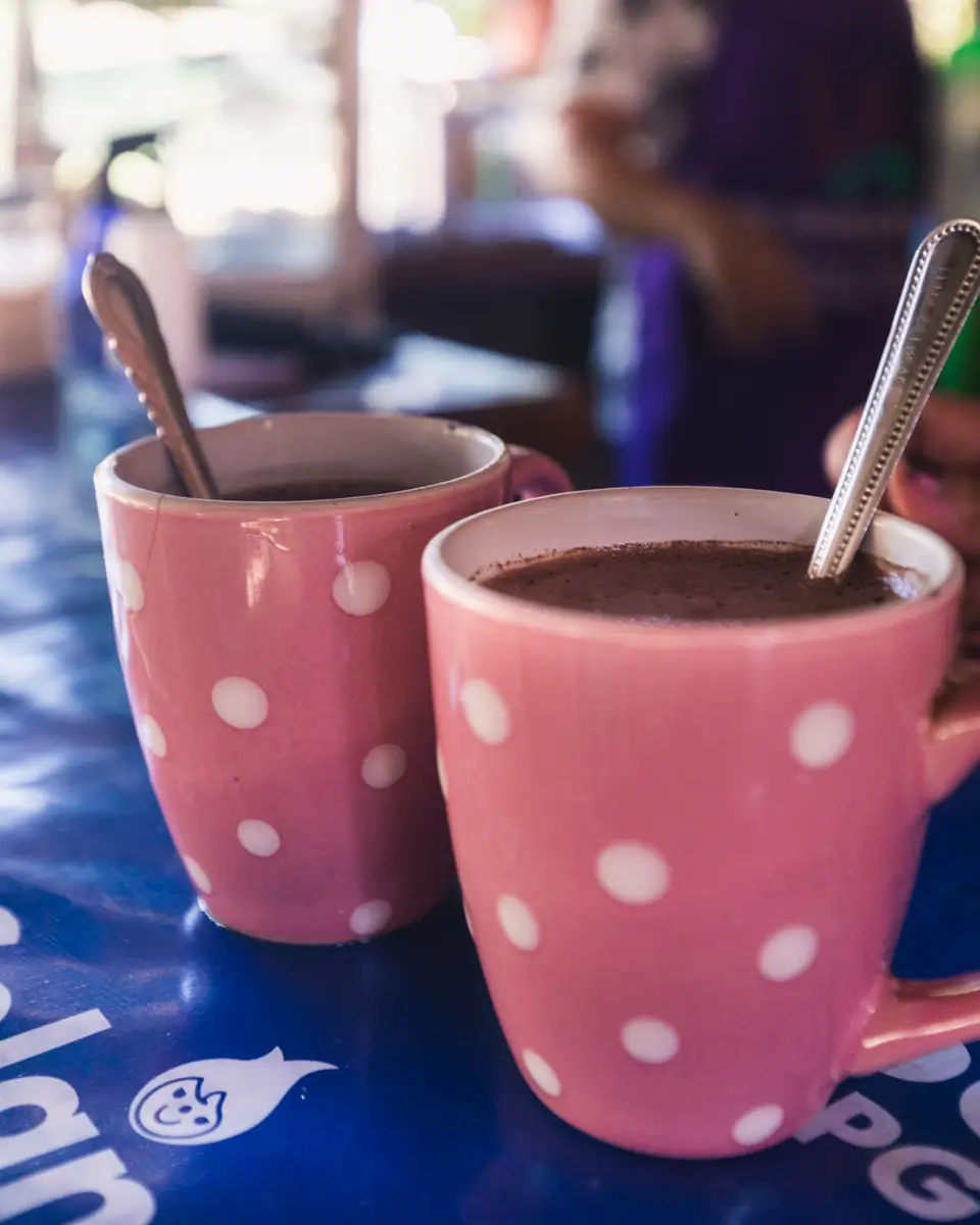 Two pink mugs with white polka dots, each containing a spoon, sit on a blue table in Bohol. The mugs appear to be filled with a hot beverage, possibly hot chocolate. The background is slightly blurred.
