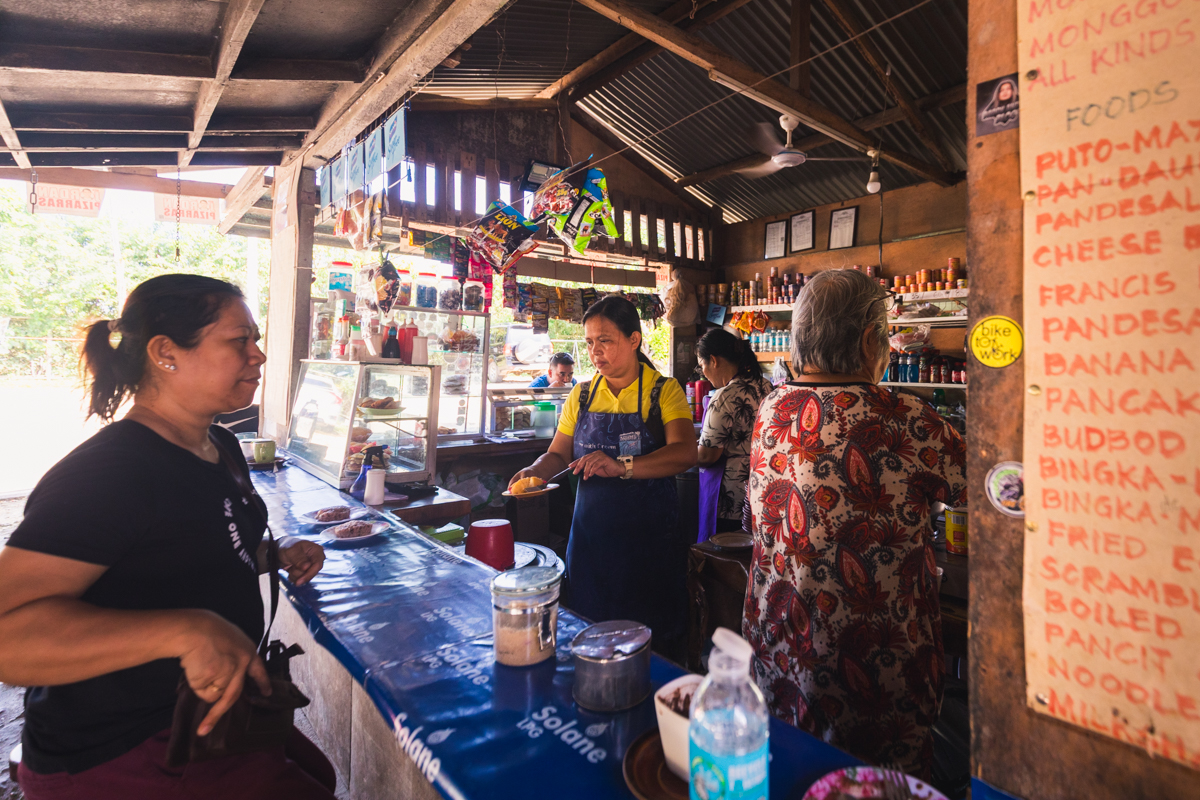 A group of people stand at the counter of a rustic Bohol eatery. A woman in a yellow shirt and apron serves food, while others wait or make purchases. A menu is displayed on the right side of the image.