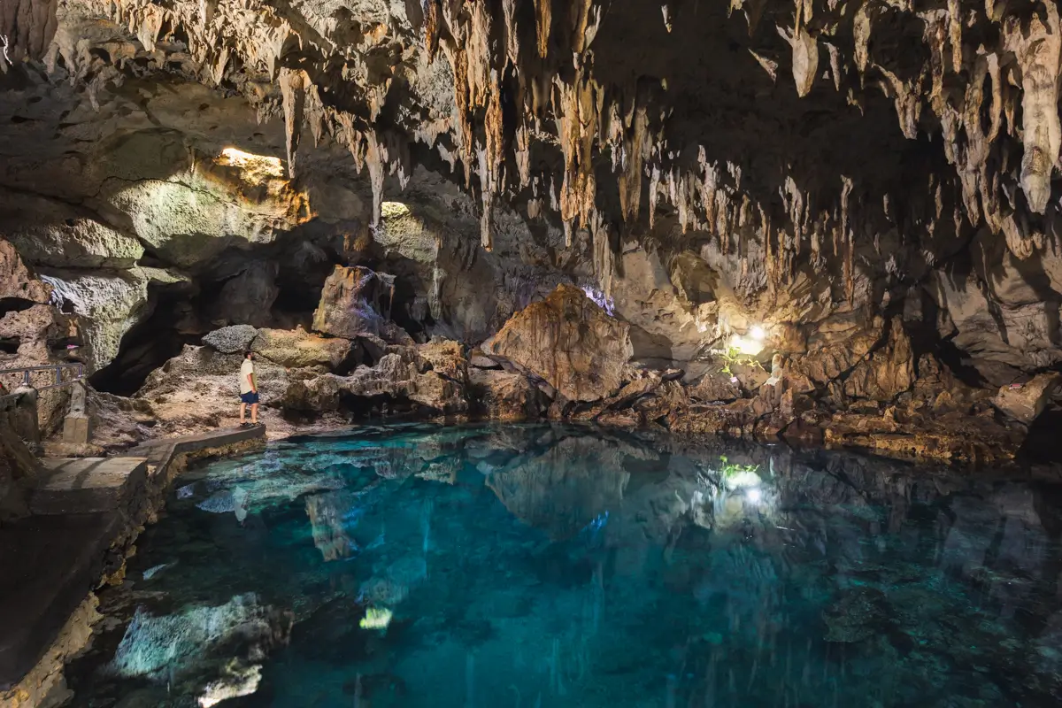 A person stands near the edge of a clear blue underground pool inside a cave in Bohol, surrounded by jagged rock formations and hanging stalactites, with electric lights illuminating parts of the scene.