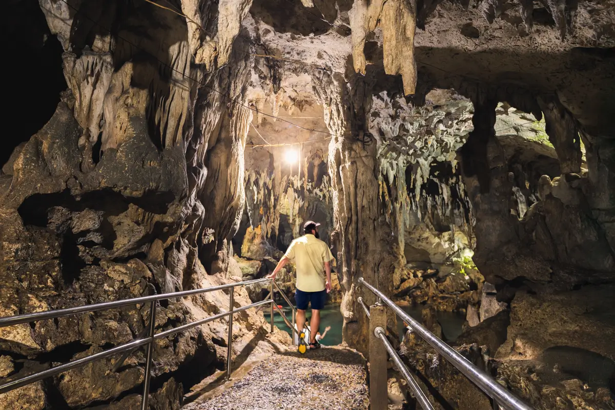 A person in a yellow shirt and shorts walks on a gravel path with metal railings inside a Bohol cave, where jagged rock formations and overhead lights illuminate the space.