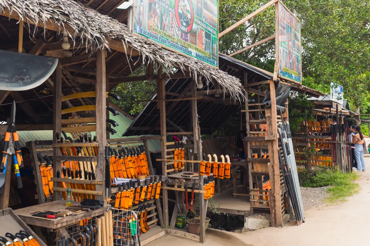 A wooden roadside shop in Bohol displays various machetes and knives with orange handles hanging on racks and shelves; two large colorful signs are posted above the shop, and trees are visible in the background.