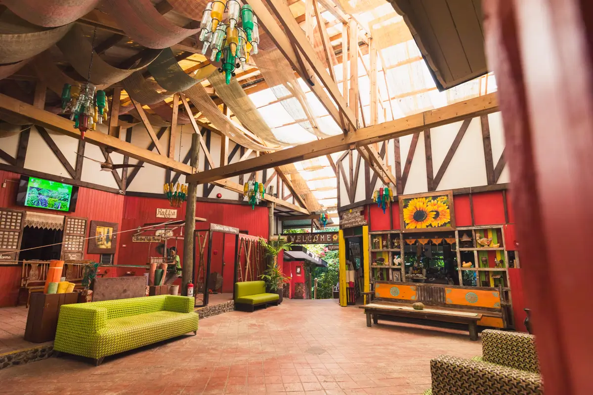 A spacious indoor area in Bohol with high wooden beams, colorful walls, green sofas, a wooden bench, potted plants, and decorative lights hanging from the ceiling. Sunlight streams through the partially transparent roof.