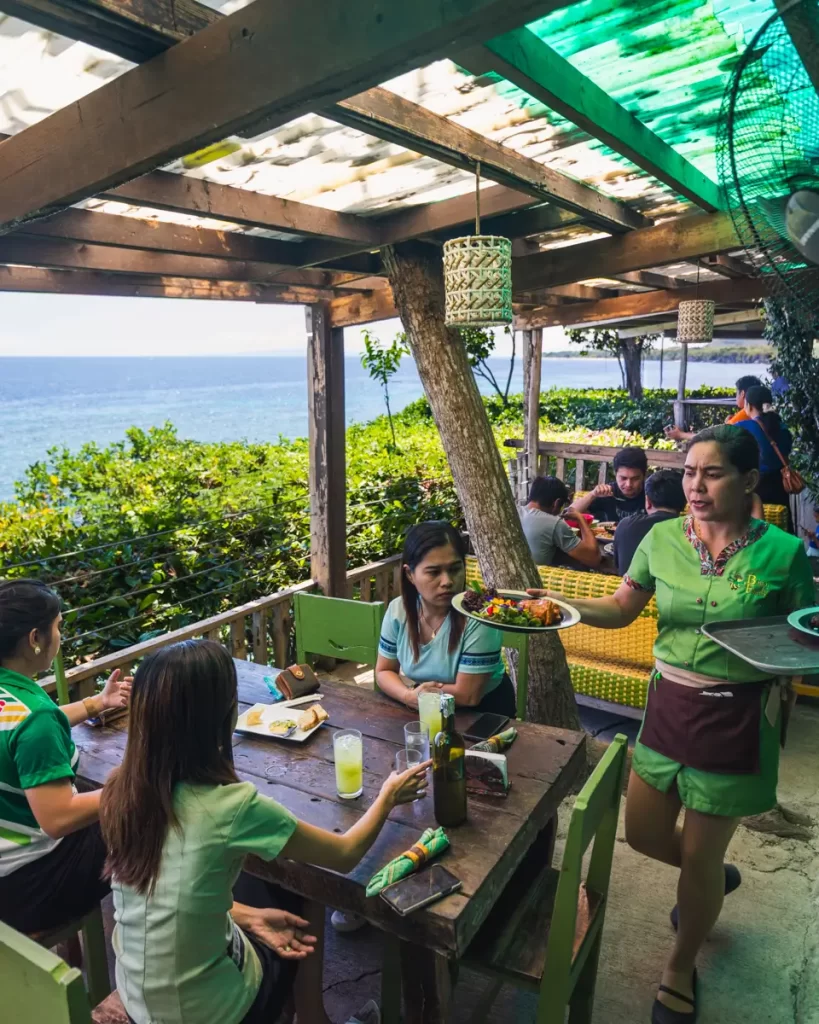 A waitress carries a tray of food to a group of three people seated at an outdoor wooden table overlooking the ocean in Bohol, under a covered patio with lush green plants surrounding the area.