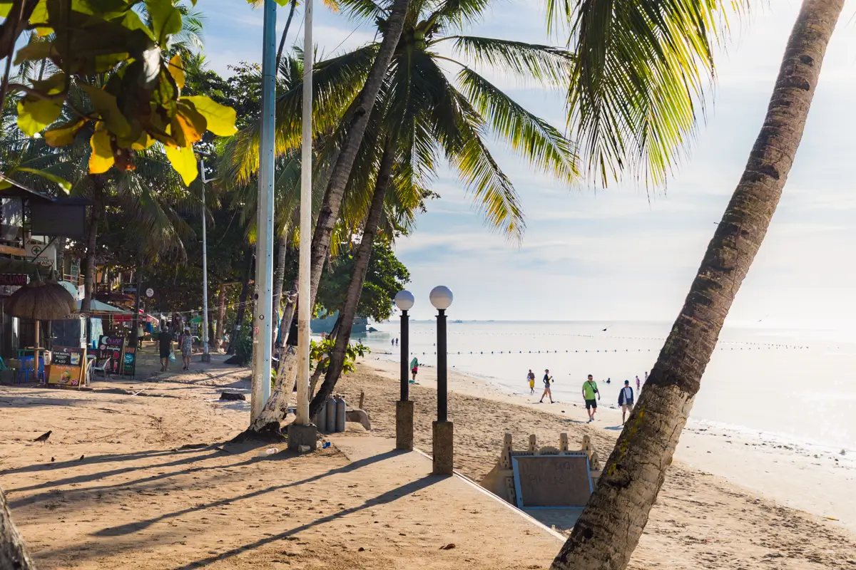 Palm trees line a sandy Bohol beach with a sidewalk, lampposts, and people strolling along the shore. Shops and buildings sit to the left, while calm sea water and a clear sky complete this scenic view.