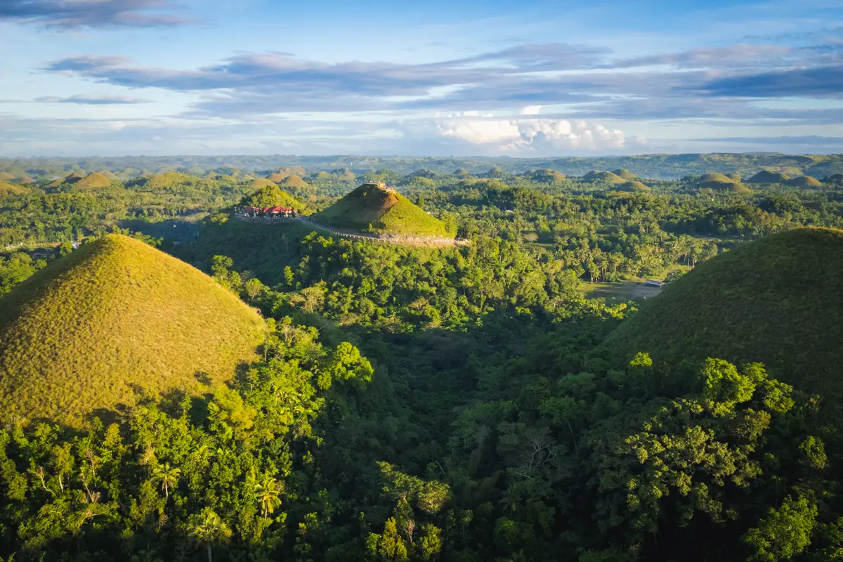 Aerial view of Bohol’s rounded, grass-covered hills surrounded by dense green forest under a blue sky with scattered clouds. A small building and pathway are visible on one of the hills.