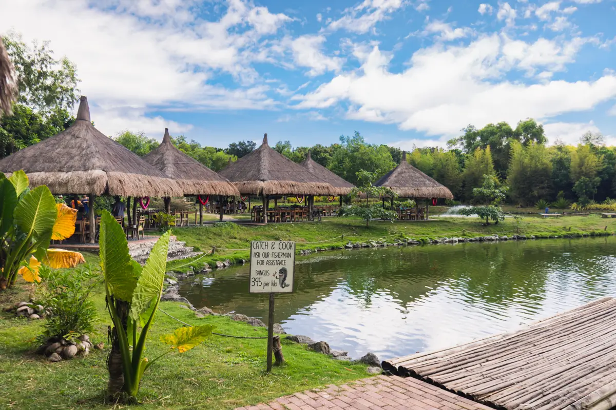 Thatched-roof huts in Bohol line the edge of a pond surrounded by greenery under a partly cloudy sky; a wooden sign with instructions and a wooden footbridge are visible in the foreground.