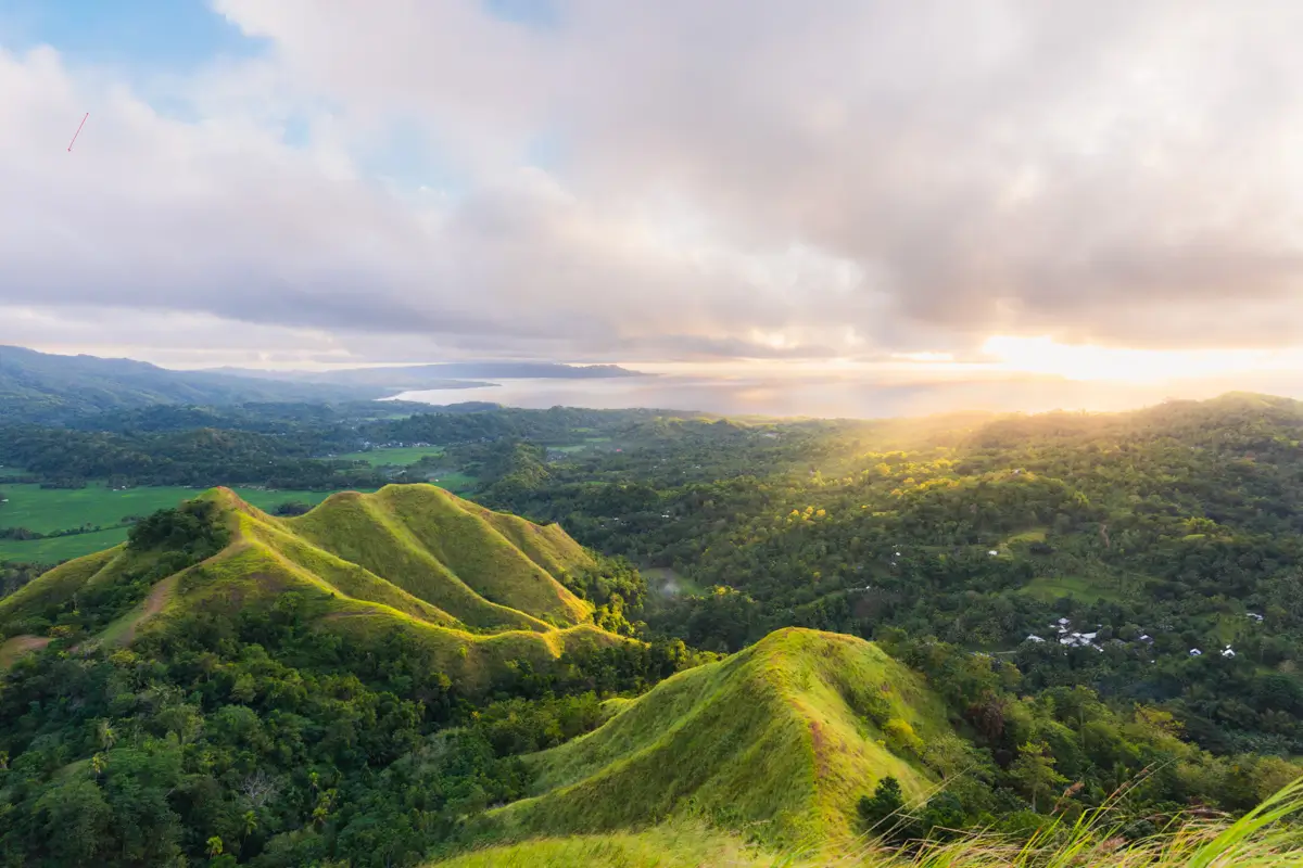 A landscape view of Bohol's green rolling hills and dense forests under a cloudy sky at sunrise or sunset, with sunlight streaming over the horizon and a distant body of water.