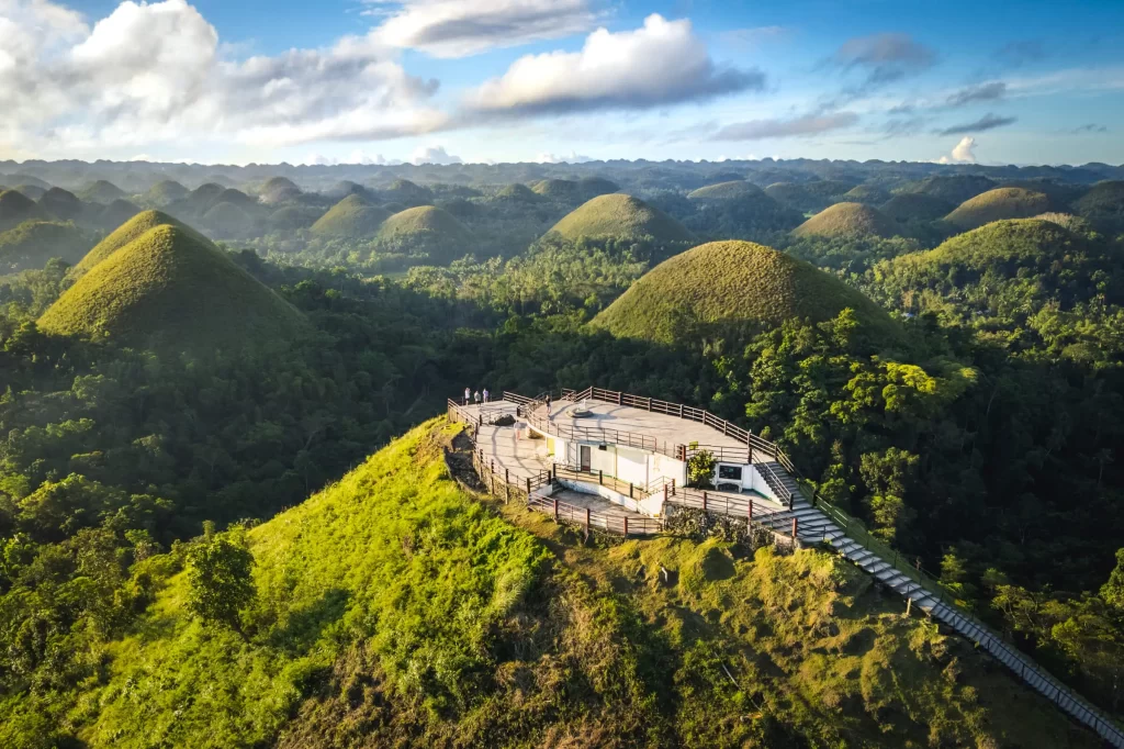 Aerial view of a hilltop observation deck in Bohol, surrounded by the Chocolate Hills—a series of green, grass-covered mounds stretching into the distance under a partly cloudy sky.
