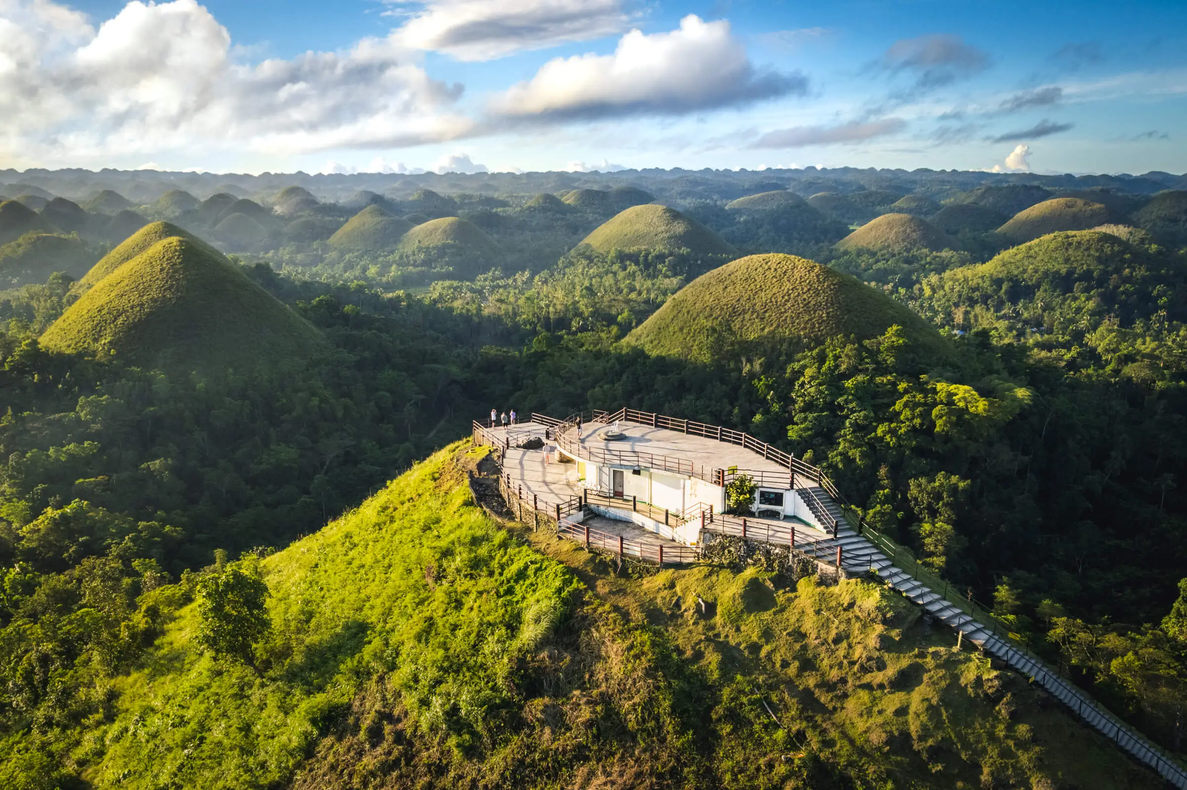 Aerial view of a hilltop observation deck in Bohol, surrounded by the Chocolate Hills—a series of green, grass-covered mounds stretching into the distance under a partly cloudy sky.