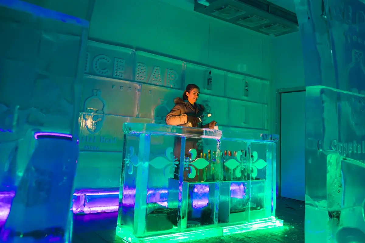 A person wearing a winter coat stands behind a bar counter made of ice blocks at Fernie, with bottles on the counter, inside a room lit with green and blue lights and adorned with ice sculptures and signage.