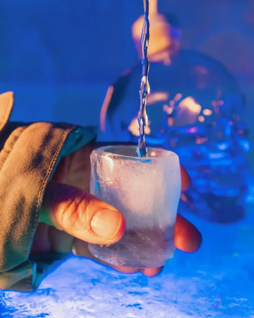 A hand holds a shot glass made of ice while liquid is poured in, with a Fernie bottle shaped like a skull in the background, all under blue and purple lighting.
