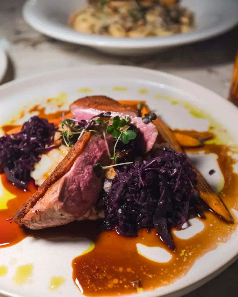 A plated dish in Fernie features sliced duck breast with a pink center, served with braised red cabbage, carrots, microgreens, and brown sauce on a white plate. Another blurred dish appears in the background.