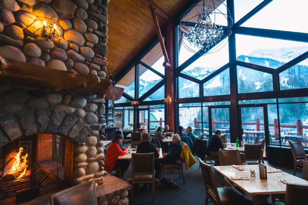 People are dining in a cozy Fernie restaurant with large glass windows showing a snowy mountain view. A stone fireplace with a lit fire is on the left, warming tables of diners arranged throughout the inviting interior.