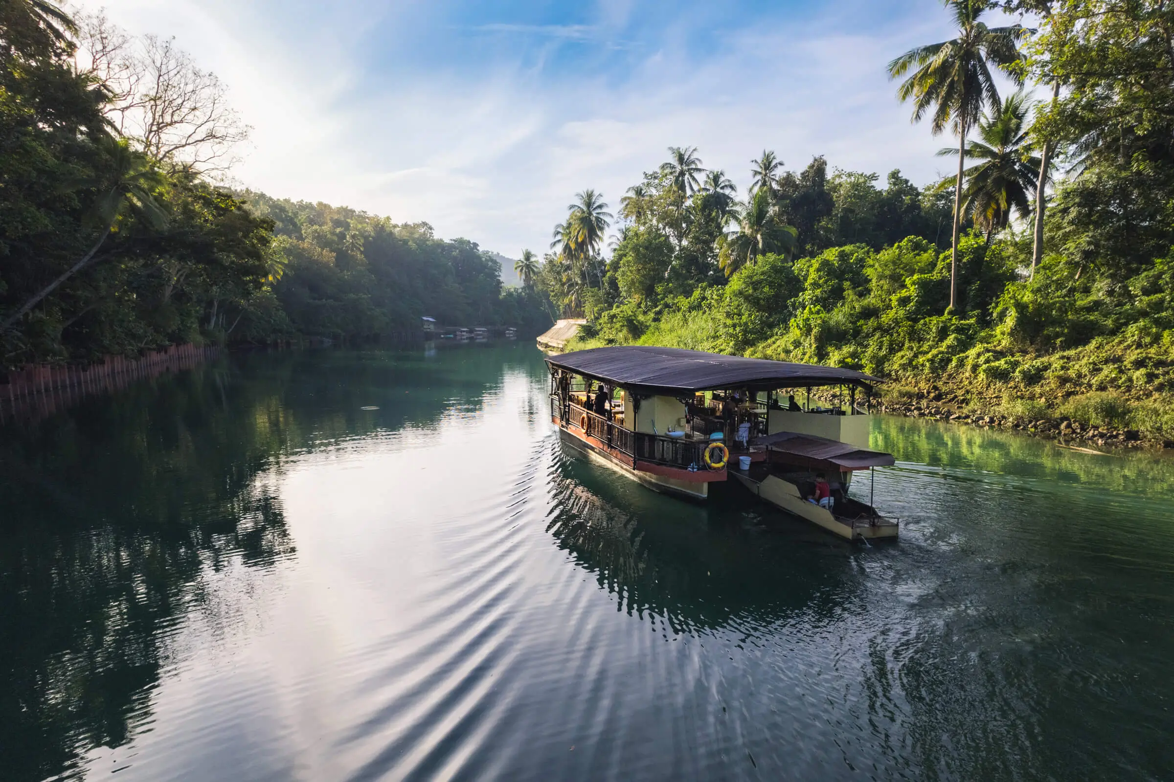 A boat with a canopy glides along a calm, green river surrounded by lush tropical trees—an idyllic scene perfect for Philippines travel, especially for first-timers seeking peaceful landscapes under a blue sky with scattered clouds.