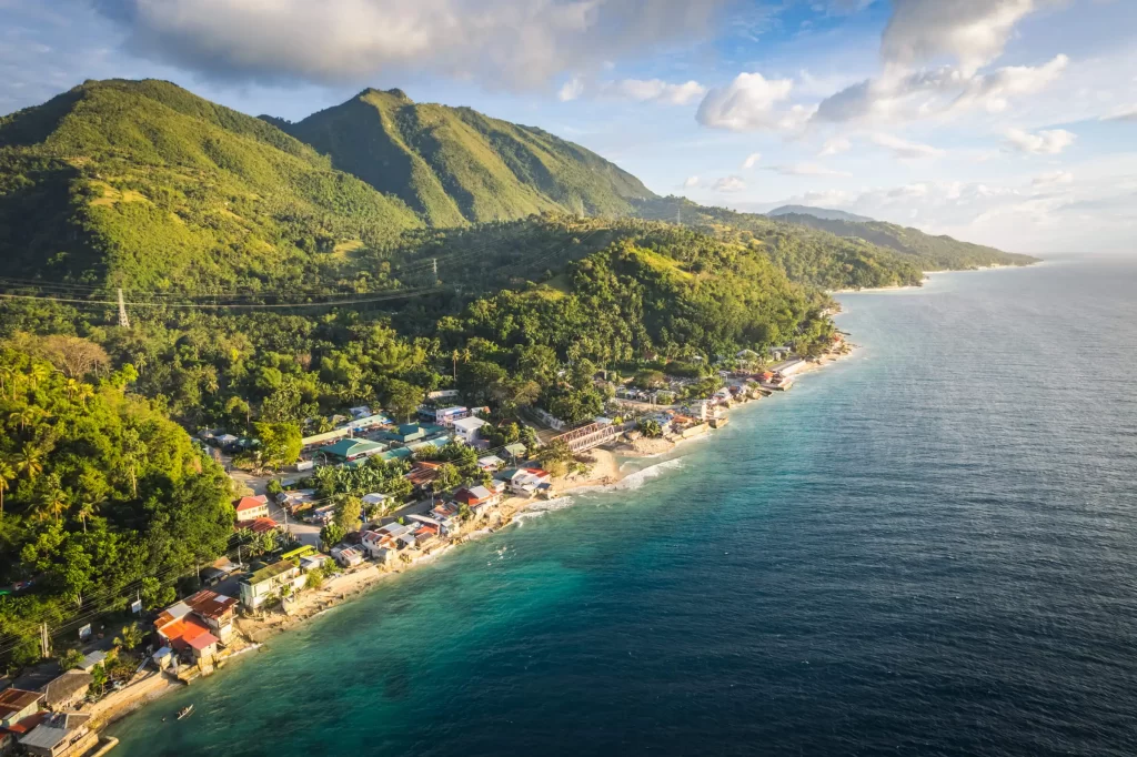 Aerial view of a coastal village with houses along the shoreline, lush green hills behind, and clear blue ocean below a partly cloudy sky—perfect for a Philippines Weather travel guide or tips on the best time to visit the Philippines.