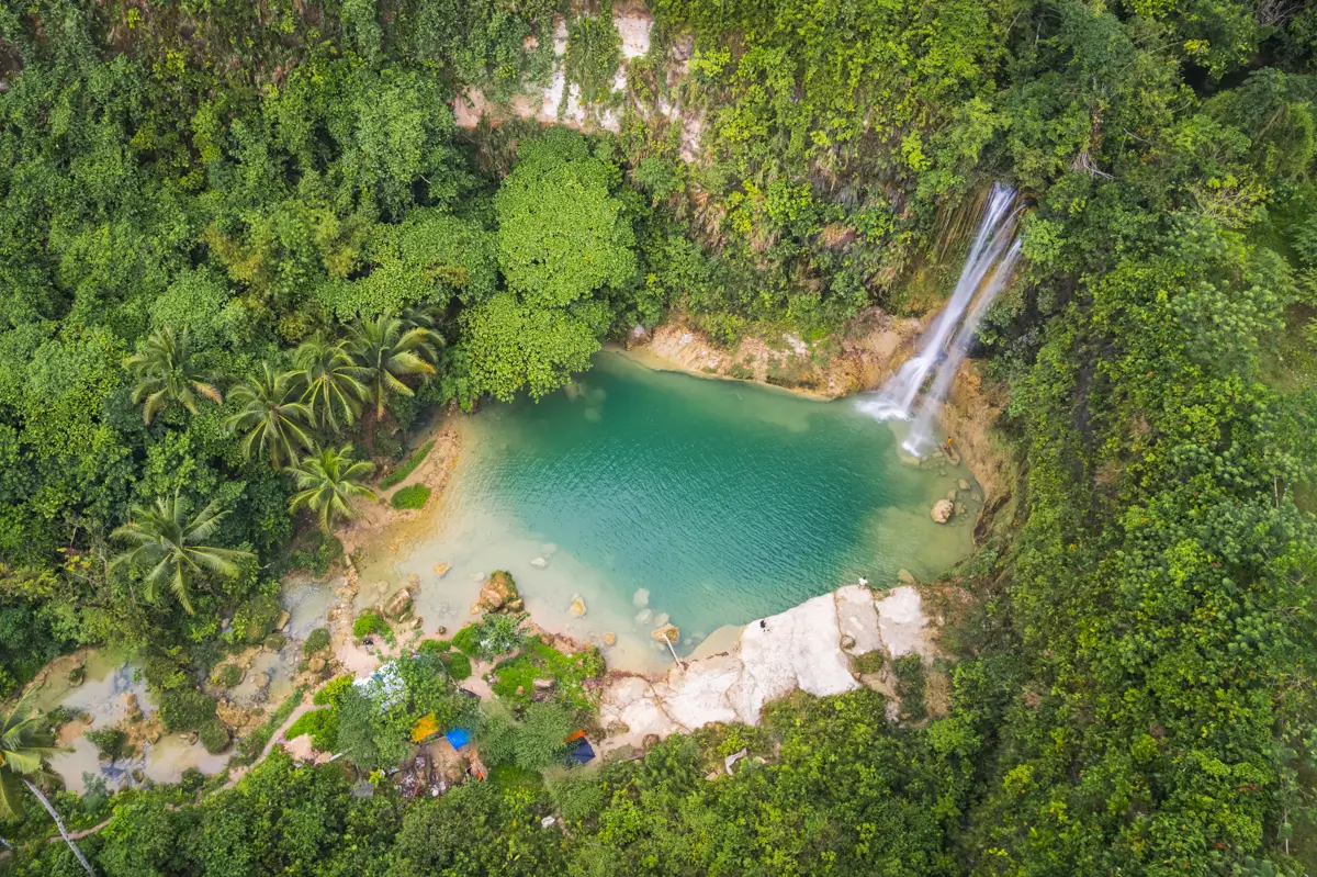 Aerial view of a turquoise waterfall pool in Bohol, surrounded by dense green forest, with water cascading from a rocky cliff into the pool and scattered rocks visible near the water’s edge.
