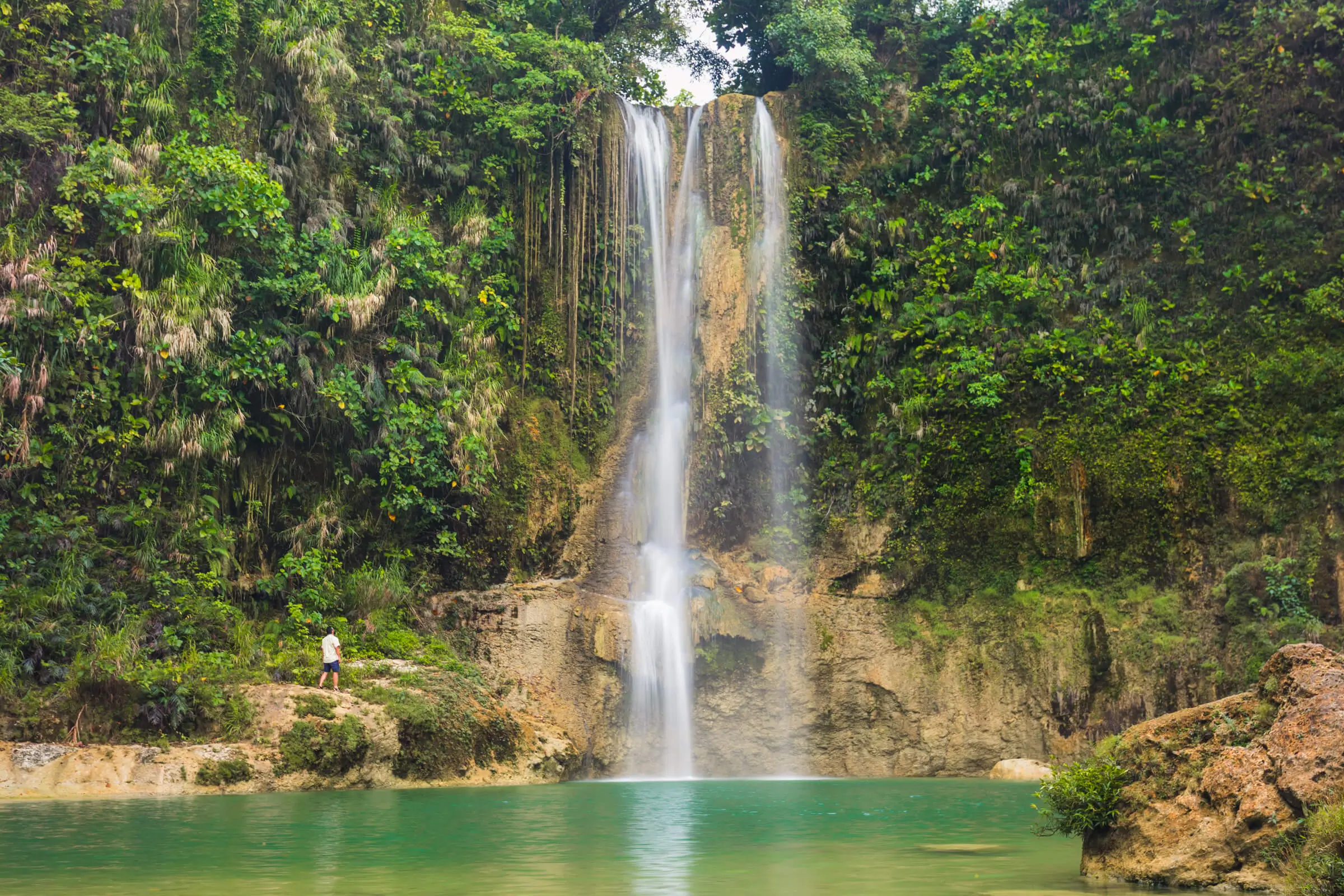 A tall waterfall cascades over a rocky cliff surrounded by dense green vegetation in Bohol, falling into a turquoise pool below. A person stands near the edge of the water, looking up at the magnificent waterfall.