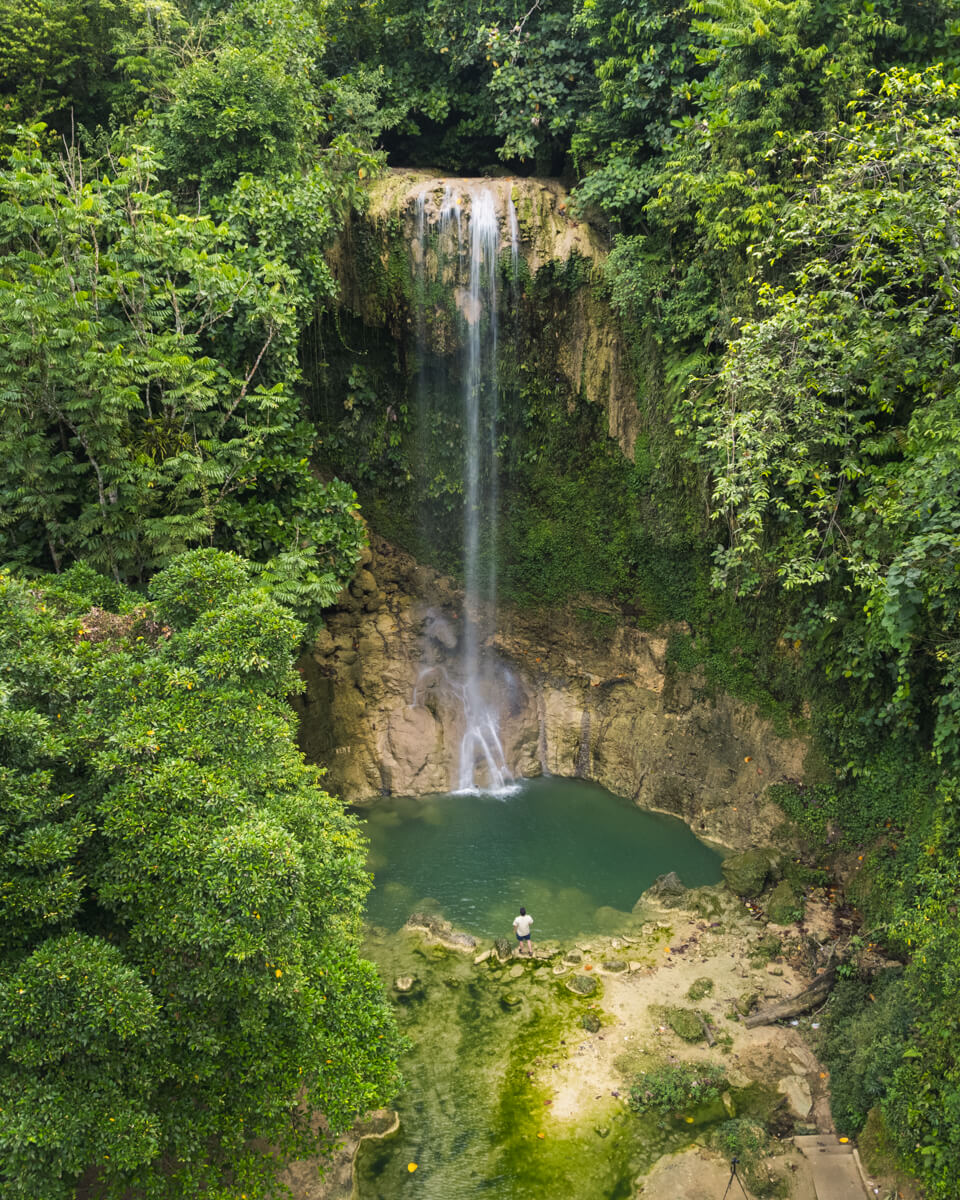 A tall, narrow waterfall in Bohol cascades into a round, green-blue pool surrounded by dense, lush rainforest. A person stands at the water’s edge, taking in the stunning scene of rocks and vibrant vegetation all around.