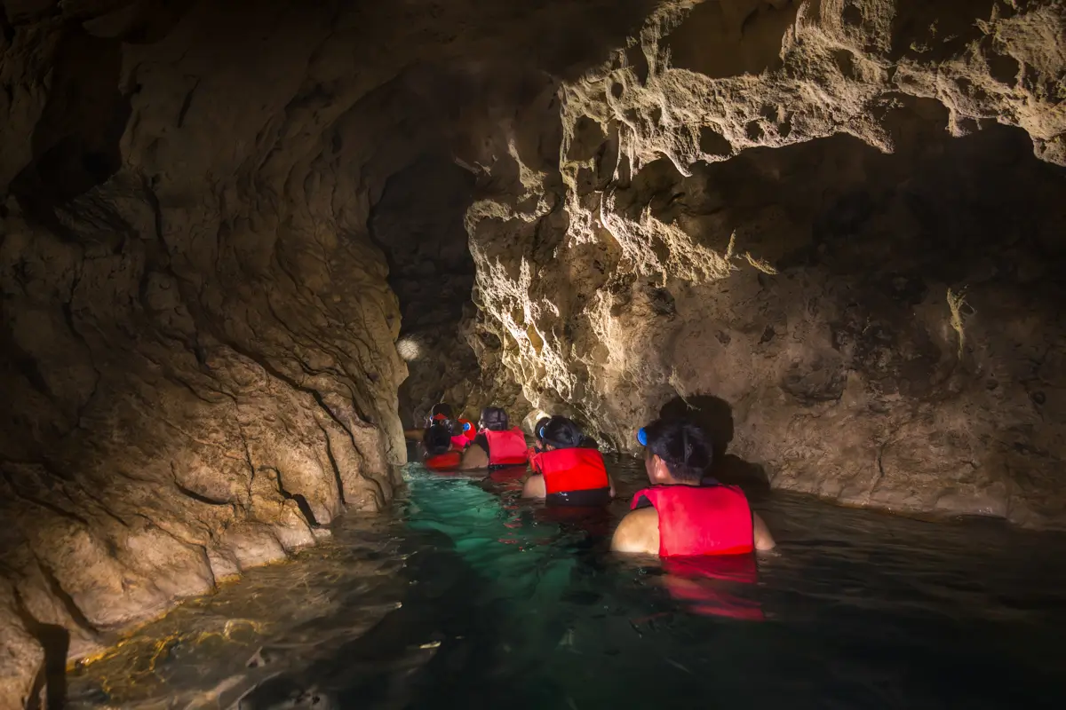 A group of people wearing red life jackets and headlamps swim through a narrow, rocky cave passage partially filled with water in Siquijor.
