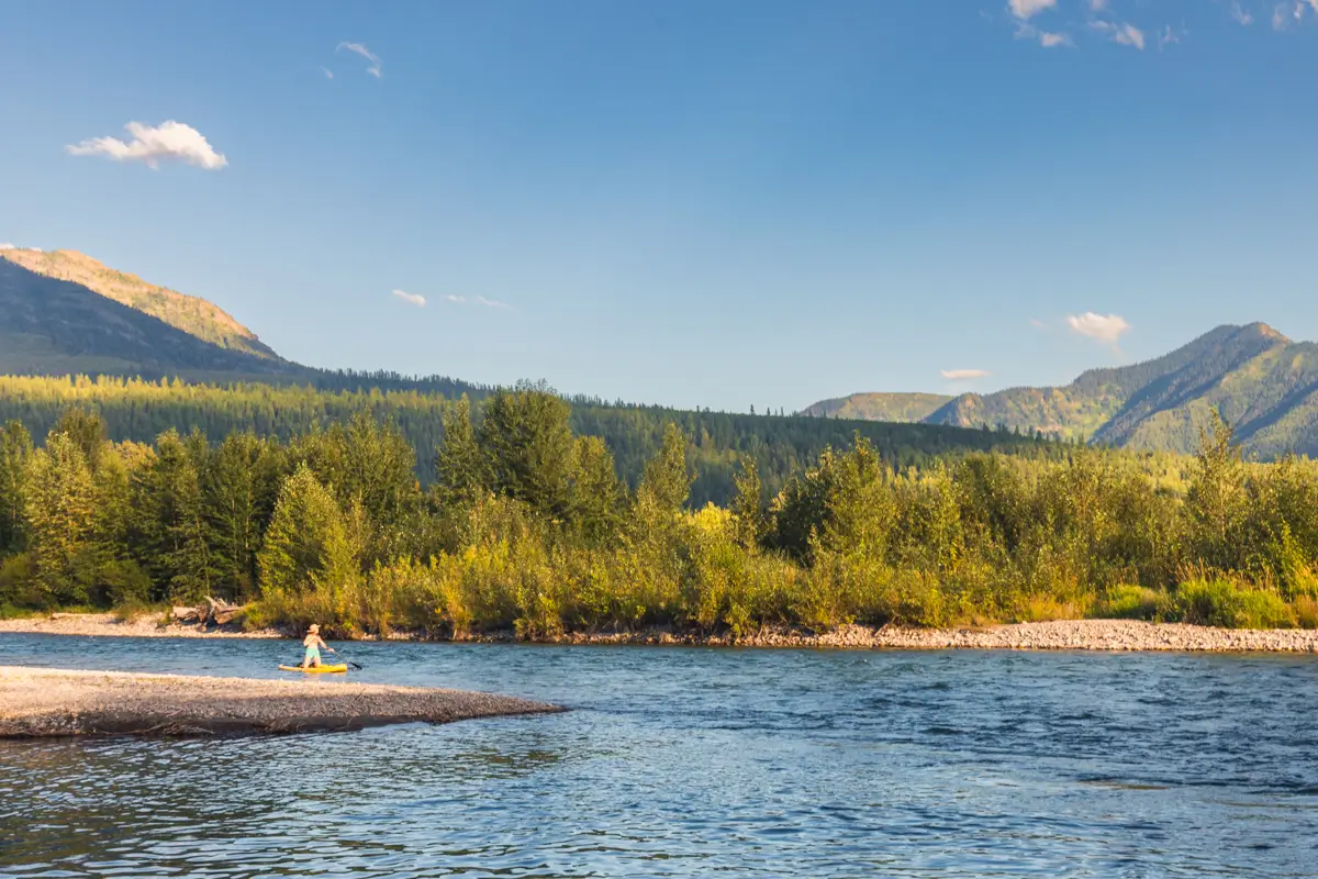A person sits on a rocky riverbank in Fernie, surrounded by water, with green trees and forested hills in the background under a clear blue sky.