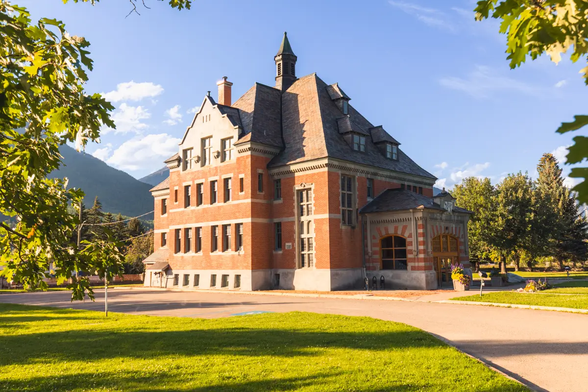 A large, historic brick building with tall windows and a steep slate roof stands on a green lawn in Fernie, surrounded by trees, under a clear blue sky in daylight.