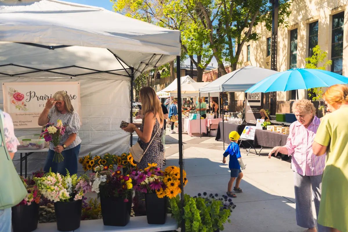 People browse flower stalls and other vendor booths at the Lethbridge outdoor farmers market on a sunny day. Several tents, tables with items for sale, and colorful flower arrangements are visible, with trees and buildings in the background.