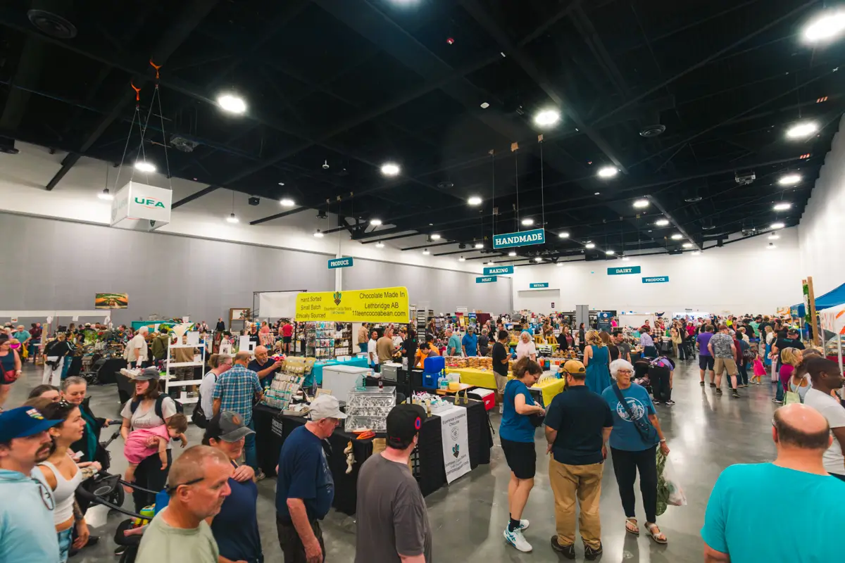 A large crowd of people walks and shops among vendor booths inside a spacious, well-lit Lethbridge convention hall. Signs above indicate sections such as “Handmade.” Various products are displayed on tables throughout the venue.