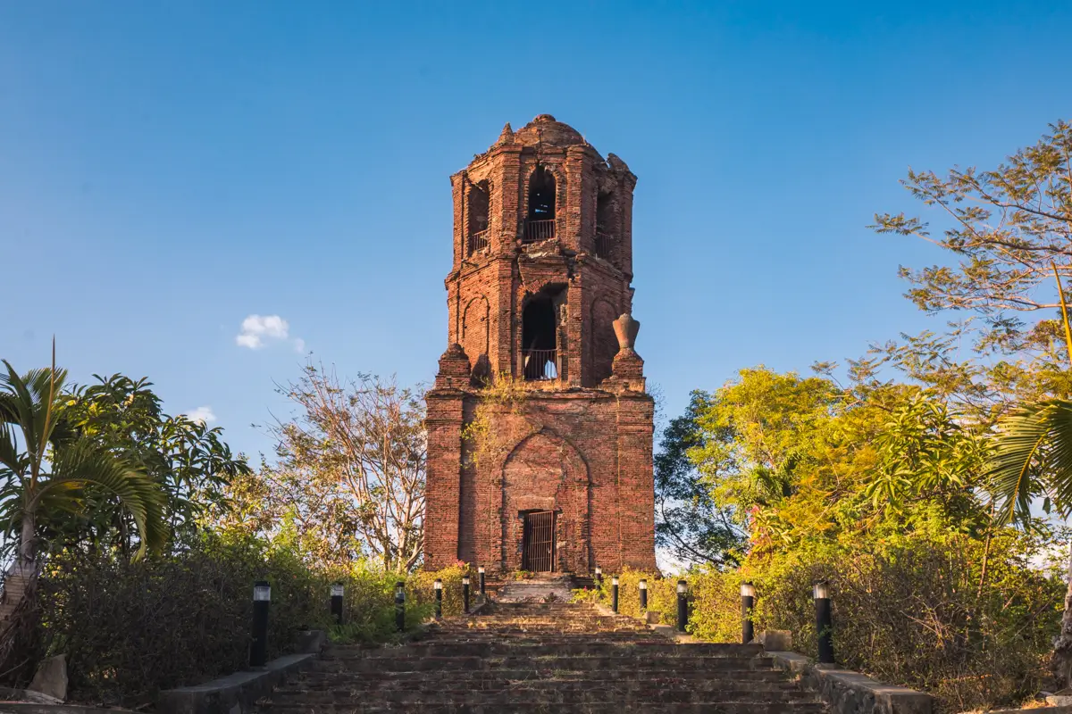 A brick bell tower with missing sections stands on a stone staircase, surrounded by green trees and plants, under a clear blue sky—one of the best places in Vigan for travelers seeking iconic Vigan tourist spots.