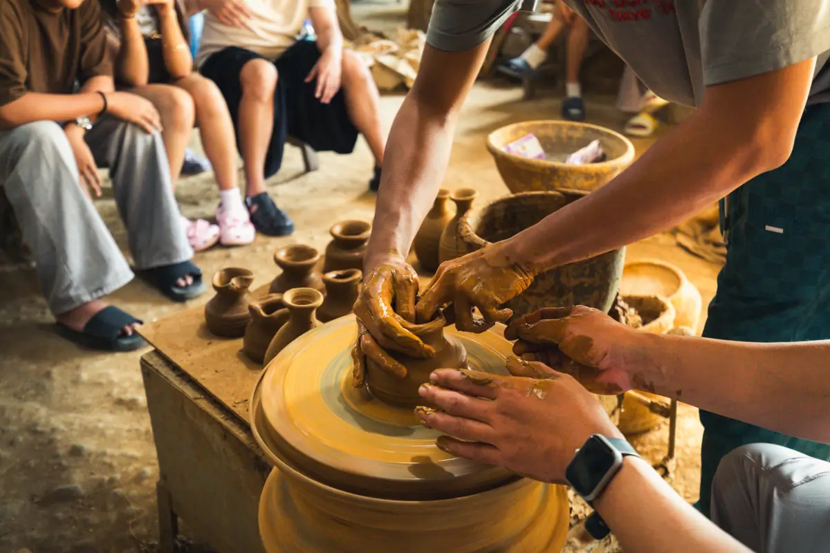 A close-up of two people shaping clay on a pottery wheel, with small finished pots and an audience in the background—a hands-on experience often highlighted in Vigan travel guide as one of the best Vigan attractions.