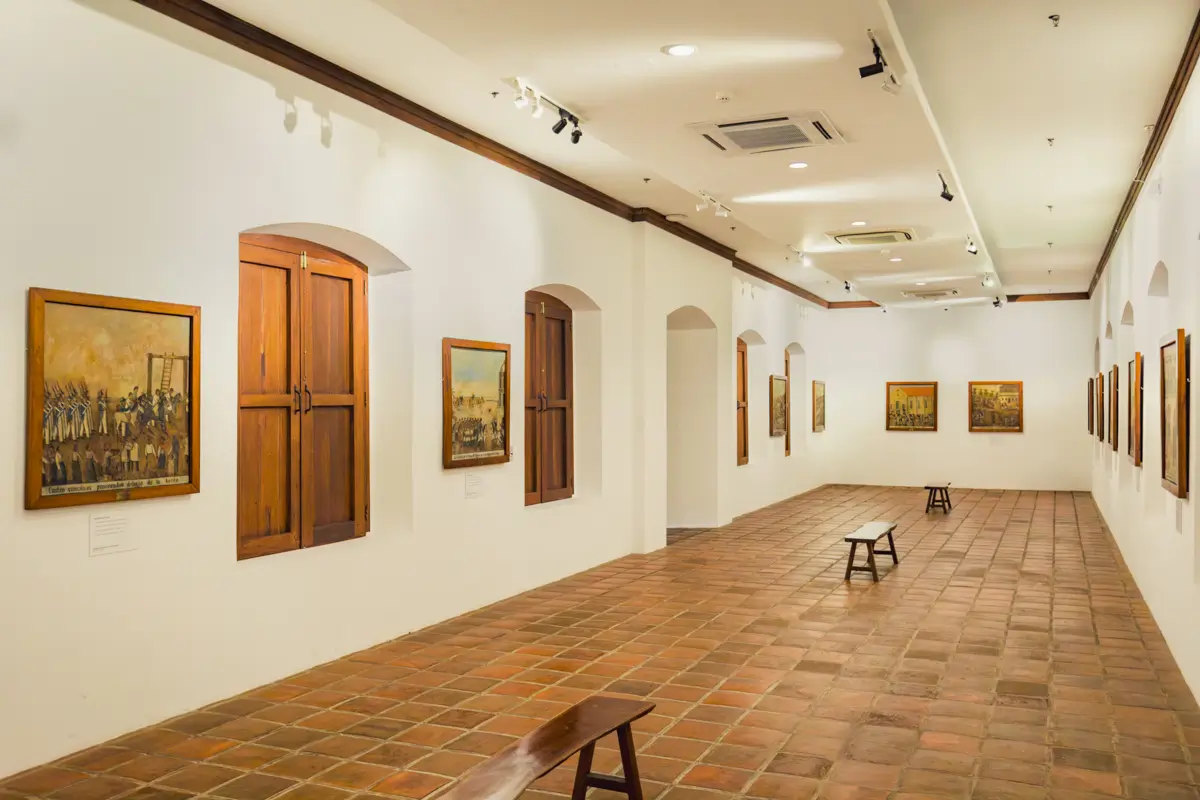 A well-lit art gallery with white walls, terracotta floor tiles, wooden benches, and framed paintings along the walls. As one of the charming Vigan attractions, it features windows with wooden shutters and ceiling lights illuminating the space.