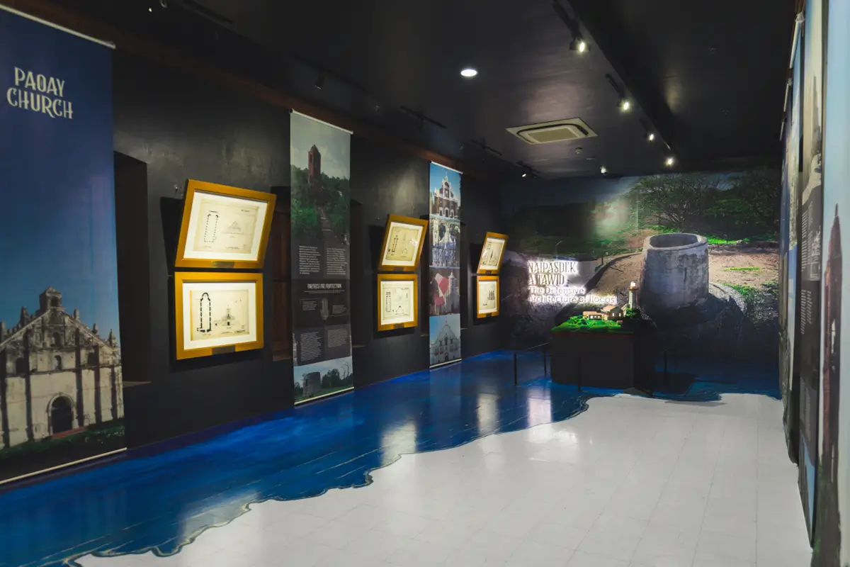 A museum exhibit room with display panels, framed documents, and photos on the walls, featuring models and historical information about Paday Church and a well—highlighting one of the best places in Vigan for history enthusiasts.