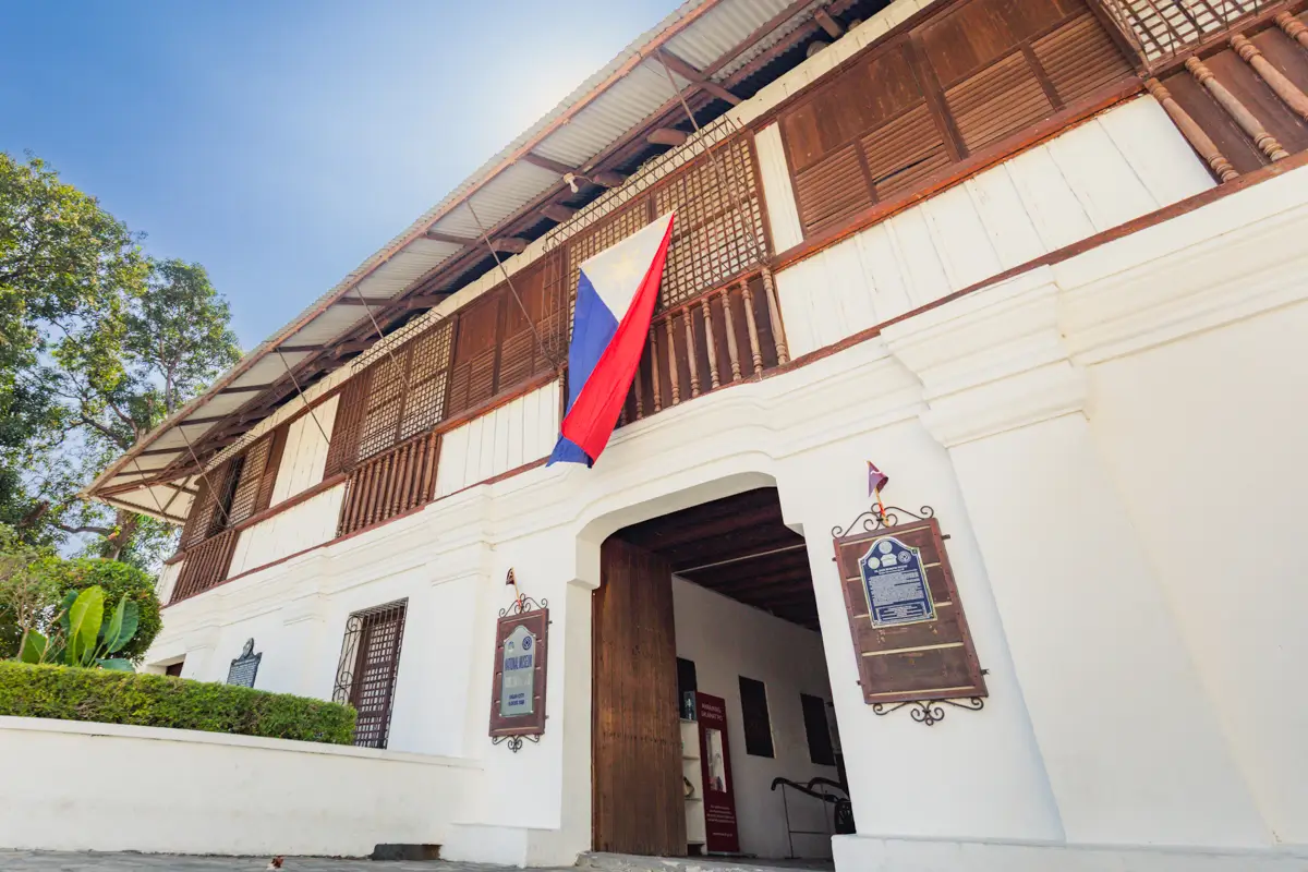 A two-story Spanish colonial-style building with white walls, wooden upper panels, a large entrance, and a Philippine flag above the doorway; plaques are mounted on either side—one of the best places in Vigan featured in many travel guides.