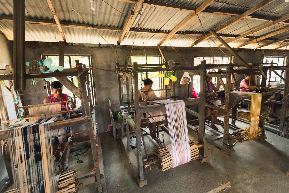 Several people operate wooden handlooms in a workshop with concrete floors and a corrugated metal roof—a fascinating scene often featured in Vigan guide lists of must-see Vigan tourist spots for those interested in traditional weaving.